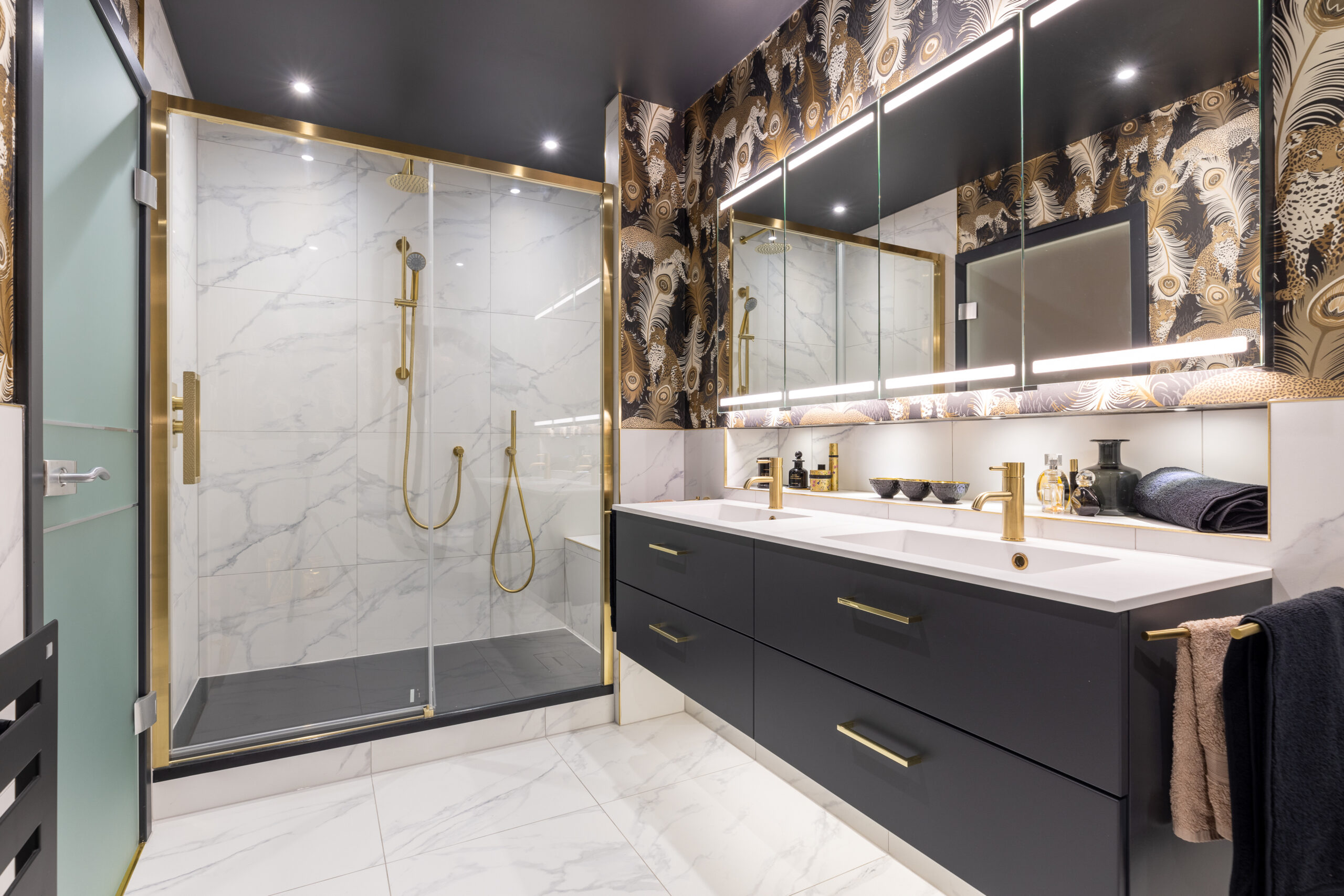 modern master ensuite bathroom with marble tile, vanity drawers, gold fixtures, dual sinks, glass shower sliding door, and wallpaper.