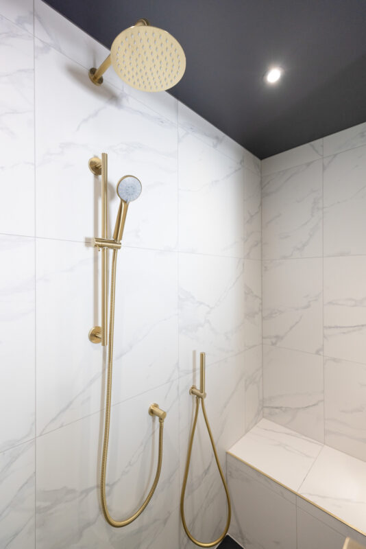 Close-up of the shower area in an ensuite bathroom featuring a large rain shower head, a handheld shower, marble-effect tiles, and a built-in bench seat.