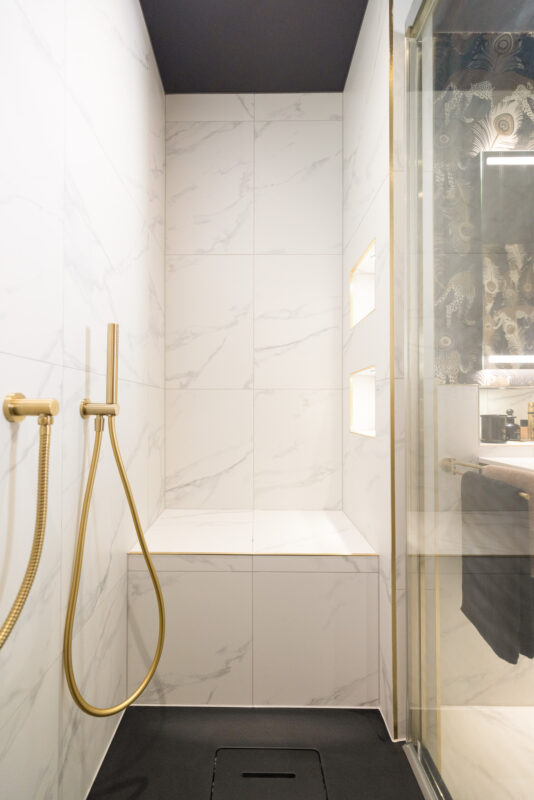 A walk-in shower with marble-effect tiles, a matte black shower tray, a built-in bench, a gold handheld showerhead, and recessed shelves in a master ensuite bathroom renovation in Edinburgh.