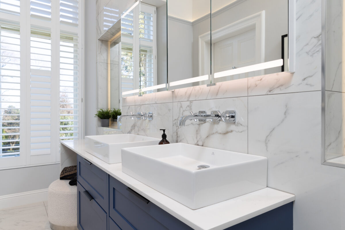 Close-up of a double sink vanity with chrome wall-mounted taps, marble wall tiles, and LED-lit mirrors in modern ensuite with steam room