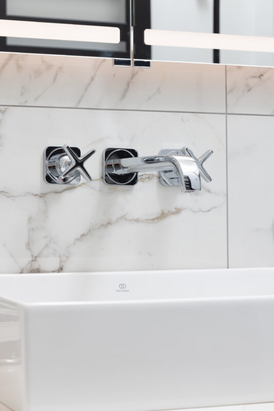 Close-up of chrome wall-mounted cross-handle taps over a rectangular white basin with marble backsplash.