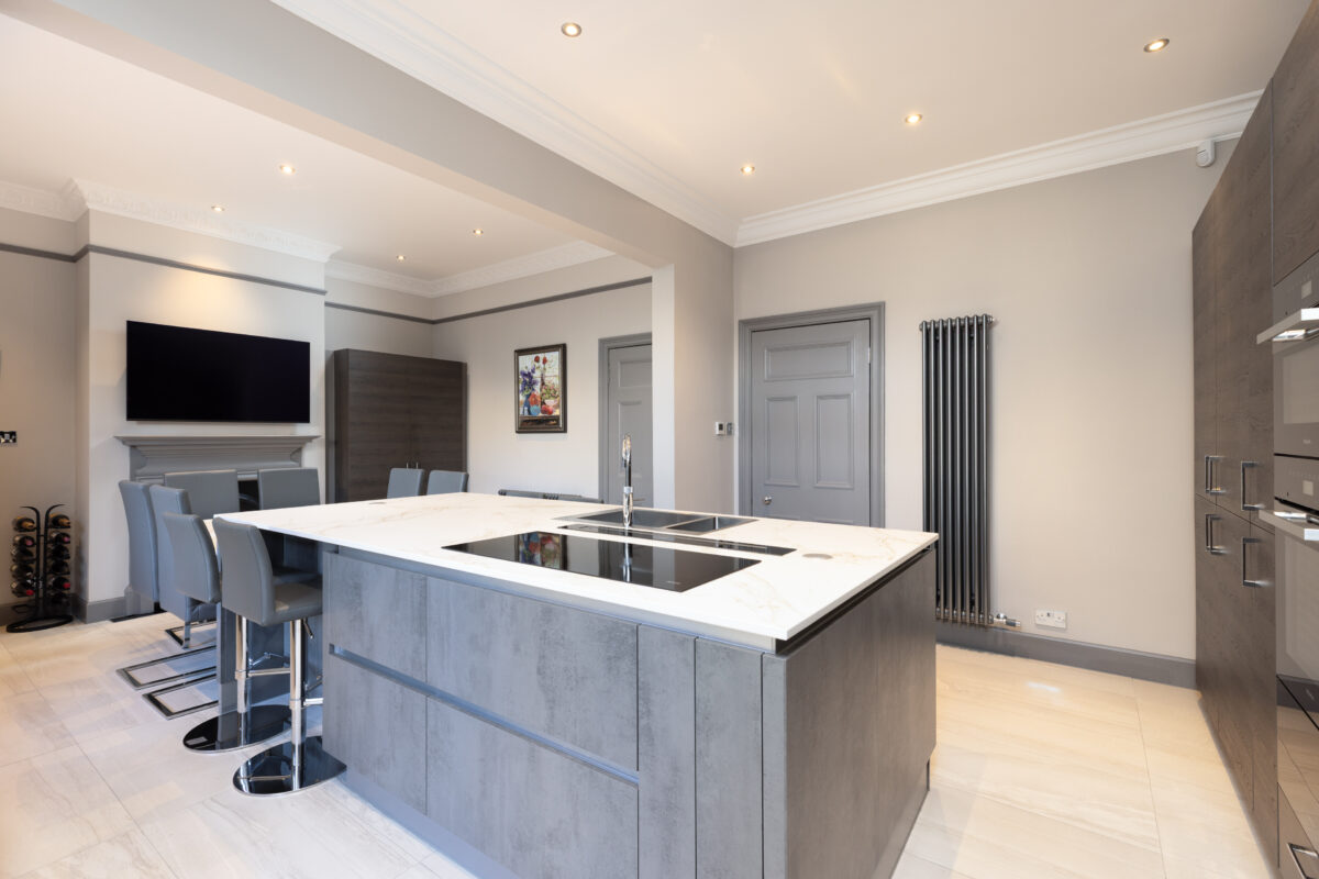 Spacious modern kitchen featuring a central Dekton countertop island, built-in induction cooktop, grey cabinets, a dining area, and a wall-mounted TV.