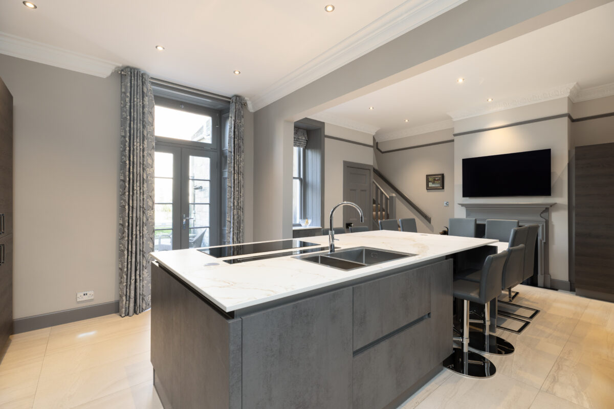 Open-plan kitchen with marble island, built-in sink and cooktop, grey leather seating, wall-mounted TV, and patterned curtains.
