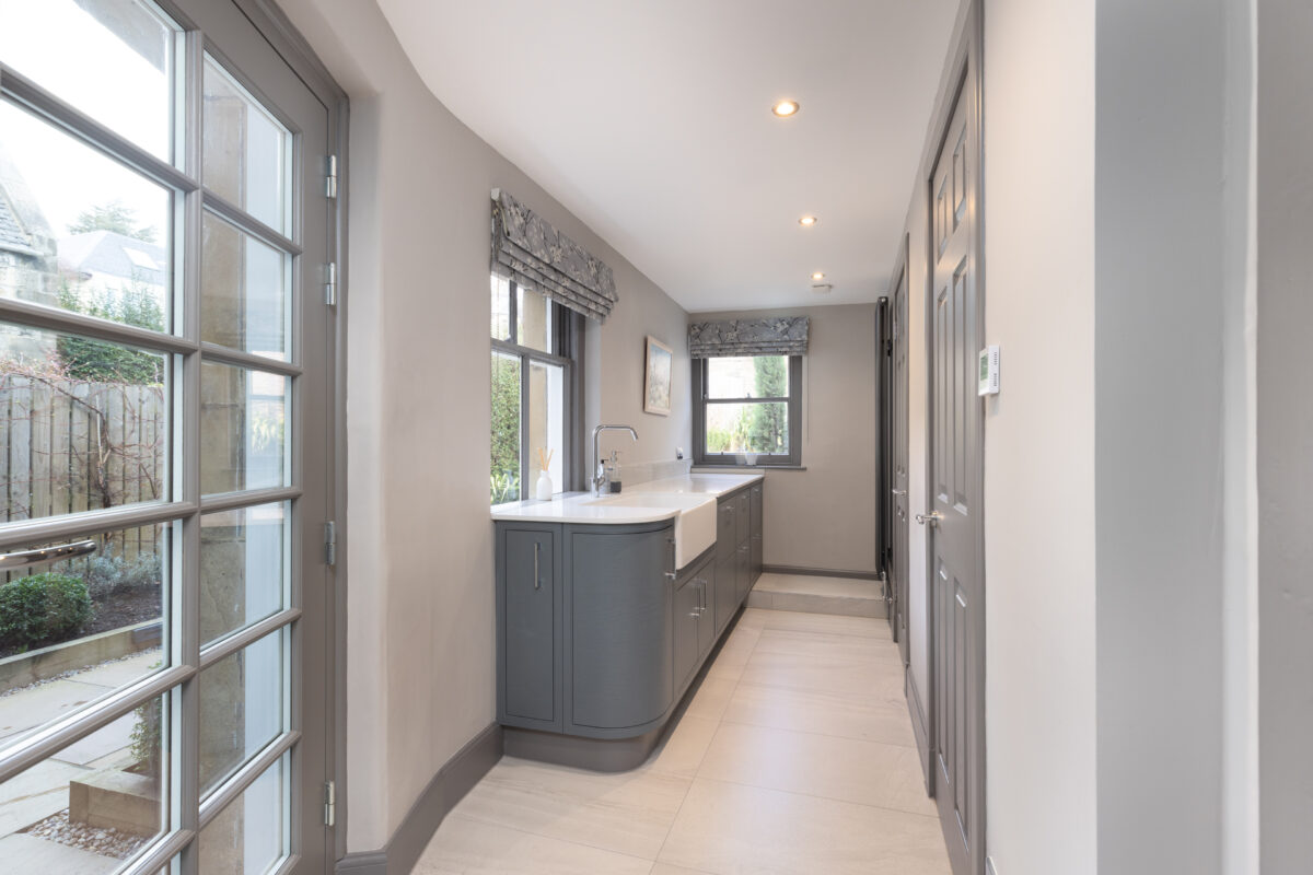 Modern utility area with grey cabinets, a white farmhouse sink, and large windows overlooking a garden as part of a full home renovation.