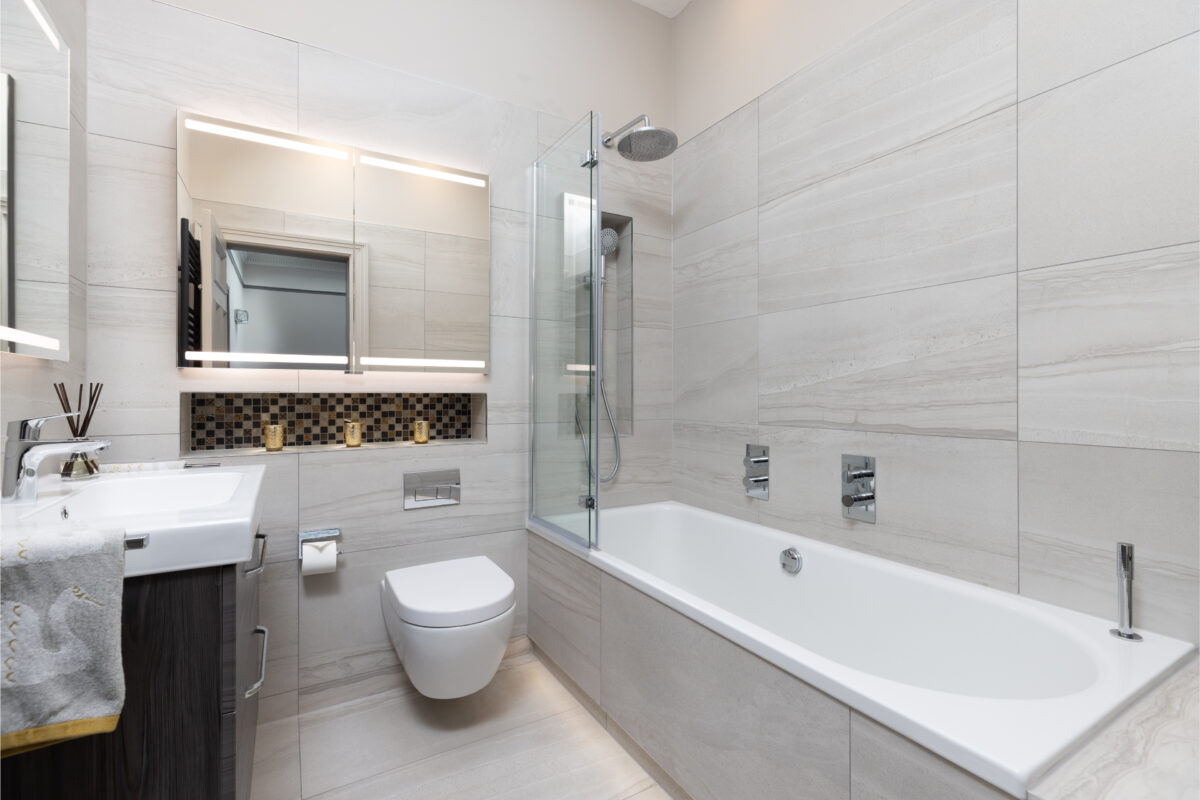 Modern bathroom with beige stone tiles, wall-mounted toilet, bathtub with glass shower screen, illuminated mirror cabinet, and mosaic shelf with gold candles that completes a whole home renovation in Edinburgh