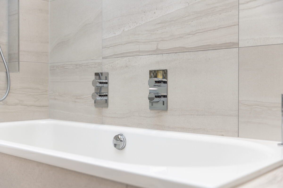 Close-up of modern white bathtub with chrome wall-mounted fixture and fittings against beige stone tile wall.