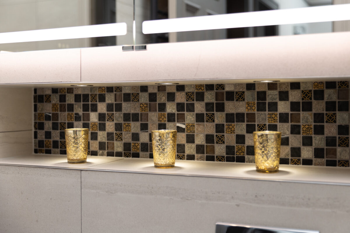 Backlit bathroom recess with mosaic tile and three gold textured candle holders under soft spotlights.