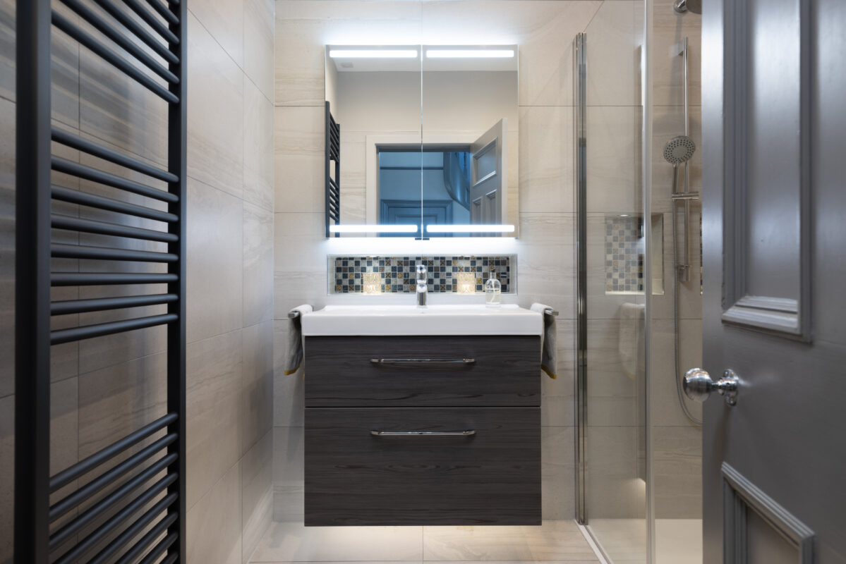 Modern bathroom with floating vanity, illuminated mirror, mosaic tile recess, walk-in shower, and matte black towel radiator in a newly renovated bathroom.