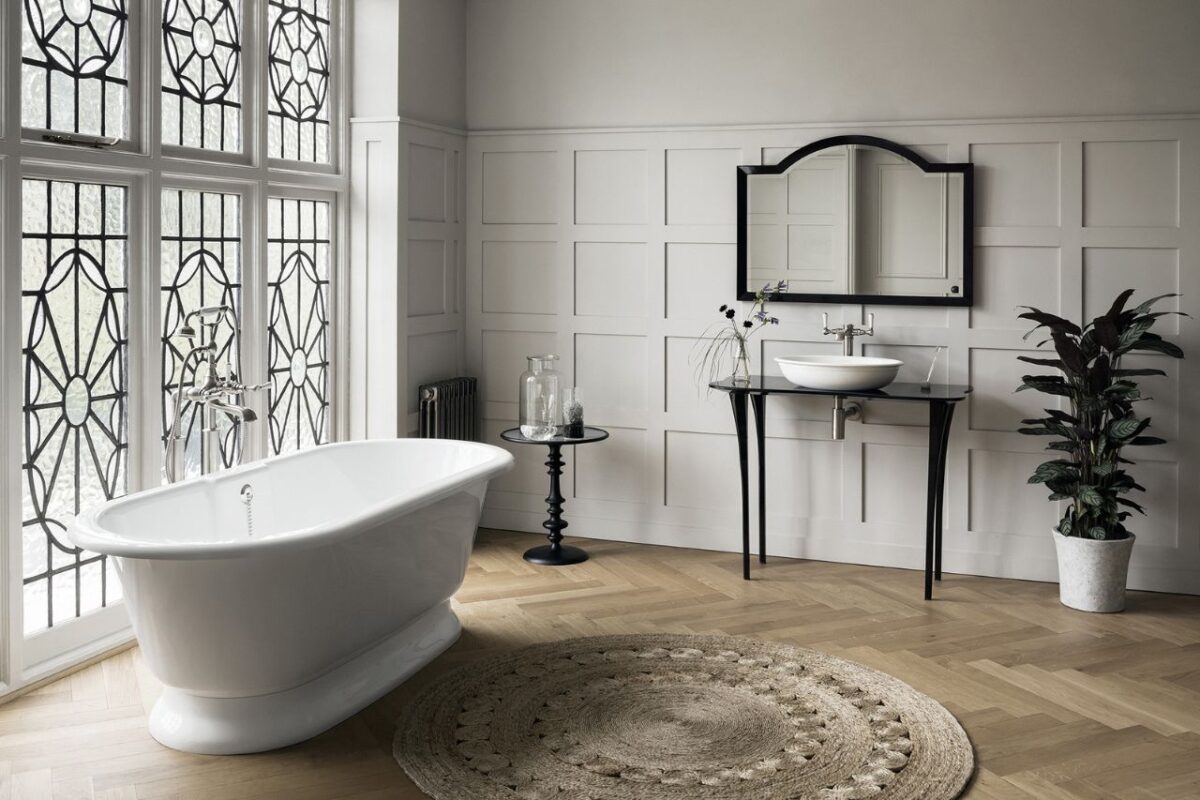A large traditional bathroom features a freestanding bath. The bath is positioned close to a large window, allowing natural light into the bathroom space. A vanity unit is adjacent to the bath area, with a mirror mounted above the vanity.