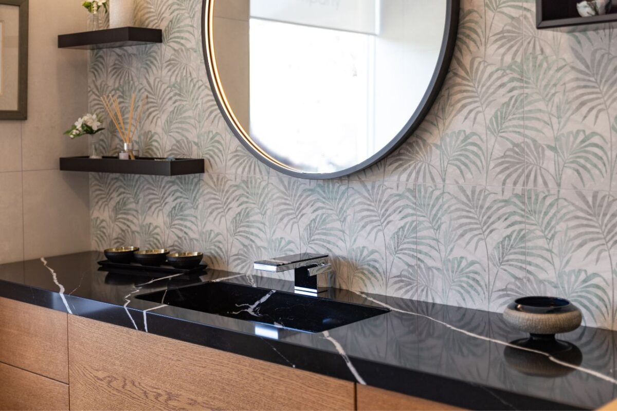 A large vanity unit with a wallpaper splashback enhancing the modern look. A large round LED mirror and floating shelves complement the space.